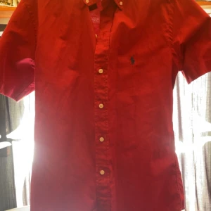 Röd kortärmad skjorta från Ralph Lauren - Säljer en röd kortärmad skjorta från Ralph Lauren med klassisk krage och knappar framtill. Skjortan har en broderad logga i blått på bröstet och är i bomull. Perfekt för en stilren och avslappnad look. Ny pris 1999kr