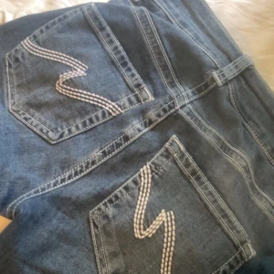 Blå jeans med broderade bakfickor från Nelly - Säljer ett par mörkblåa jeans som är bootcut och snygga broderade detaljer på bakfickorna. Jeansen har dubbla knappar i midjan och klassisk femficksdesign. Perfekta för dig som gillar en tidlös look med en twist. Säljer dom för dom är för små i midjan för mig! Använt 2-3 gånger, nytt pris: 699