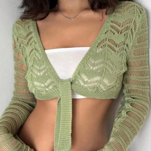 Grön stickad bolerotopp med knyt - Snygg ljusgrön stickad bolerotopp med lång ärm och knyt framtill. Toppens mönster är hålstickat i vågigt zigzag-mönster och den är croppad. Perfekt att styla över ett linne eller tubtopp.