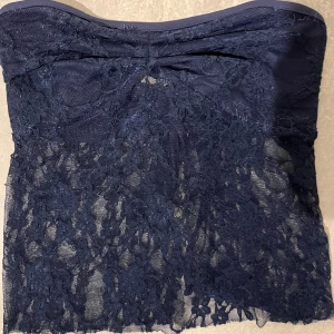 Mörkblå spets tubtopp - Säljer en mörkblå tubtopp i spets med transparenta partier och fina spetsdetaljer. Perfekt att styla till olika outfits för en trendig look. Toppen är axelbandslös och har en romantisk känsla.