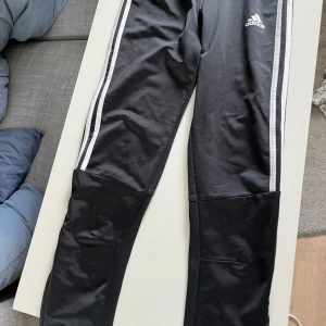 Svarta träningsbyxor från Adidas - Svarta träningsbyxor från Adidas med klassiska vita ränder längs sidorna och Adidas-logga på låret. Byxorna har dragkedja vid benslut och är tillverkade i ett syntetmaterial som passar perfekt för sport och träning.