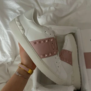 Vita sneakers med rosa detaljer från Valentino - Säljer ett par vita sneakers från Valentino med bred rosa rem över vristen dekorerad med nitar. Skorna har platt sula och rund tå, samt nitar längs bak på sulan. Perfekta för dig som gillar en stilren men edgy look.