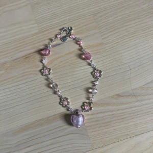 Sött armband med rosa hjärta och blommor - Armband med silverfärgade blommor, rosa pärlor och ett stort rosa hjärta som hänge. Fina detaljer och lekfull design, perfekt för att ge en gullig touch till din stil.