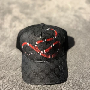 Svart keps med ormtryck från Gucci - Säljer en Svart keps från Gucci med diskret GG-mönster och en röd, vit och svart orm tryckt framtill. Kepsen har böjd skärm och justerbar rem baktill