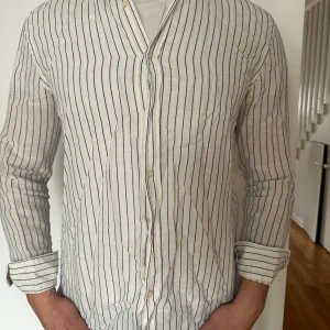 Vit och blårandig skjorta - Säljer en vit skjorta med tunna blå ränder. Skjortan har lång ärm, knappar framtill och klassisk krage. Perfekt för dig som gillar en stilren och enkel look. Materialet är i linne. Modellen är 183 cm lång.