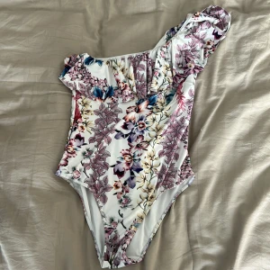 Blommig baddräkt med volang och asymmetrisk axel - Aldrig använd och är i storlek M men kan även passa S. Säljer en snygg baddräkt med blommigt mönster i lila, rosa och beige toner. Modellen har en axel och volangdetalj upptill för en trendig look. Perfekt för stranden eller poolen! 