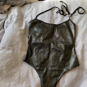 Svart body i skinnimitation - Säljer en svart body med smala axelband i skinnimitation. Snygg och stilren modell med glansig finish, perfekt att styla till olika outfits.