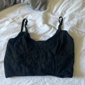 Svart spetsig linnetopp - Säljer en svart linnetopp med spetsdetaljer och justerbara axelband. Toppen har en croppad passform och är perfekt för att skapa en trendig look. Passar lika bra till jeans som till kjol.