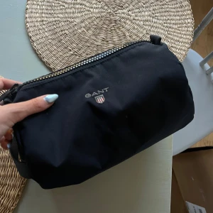 Mörkblå  necessär från GANT - Säljer en svart necessär från GANT med dragkedja och logotyp framtill. Praktisk och stilren design med ett handtag på sidan. Perfekt för att organisera dina småsaker eller ta med på resan.