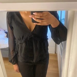 Svart blus med volang från BikBok - Säljer en svart blus från BikBok med v-ringning och volangdetaljer framtill. Blusen har långa, lätt transparenta ärmar och en lös passform. Perfekt att styla till olika tillfällen.