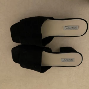 Svarta slip-in sandaler med klack från nelly - Stilrena svarta sandaler med bred rem och fyrkantig tå från nelly. Skorna har en låg klack och öppen tå, perfekta för varmare dagar. Enkla att matcha till olika outfits och lätta att ta på.