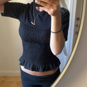 Svart smockad topp med volangkant - Säljer en svart topp med smockad midja och volangkant nertill. Toppen har korta ärmar och rund halsringning. Perfekt för dig som gillar en feminin och trendig stil.