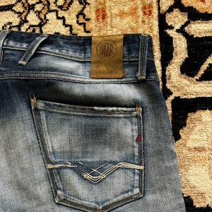 Replay Anbass jeans  - Säljer ett par Replay Anbass jeans i blå tvättad denim med snygga slitningar och klassiska fem fickor. Jeansen har normal passform och raka ben, med Replay-logga på bakfickan och läderpatch i midjan. Otroligt sällsynt och snygg tvätt. Skick 8/10. Strl 34. Pris 499