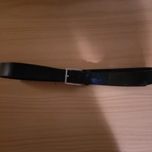 Calvin Klein svart bälte 90 cm - Ett svart bälte från Calvin Klein med ett silverfärgat spänne.Bältet har en ögla med blå detaljer och Calvin Klein -logotypen. De två skikten som bältet består av har särat på sig på två ställen (se bilder). Nypris 800 kr