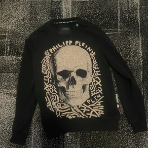 Svart tröja med dödskalle från Philipp Plein - Cool svart tröja från Philipp Plein med stort dödskalletryck framtill och grafiska detaljer runt motivet. Rund halsringning och långa ärmar. Liten PP-logga i metall bak på ryggen. Perfekt för dig som gillar statement-plagg.
