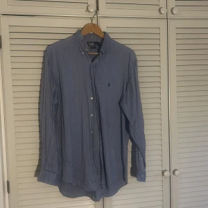 Blå skjorta från Polo Ralph Lauren - Säljer en klassisk blå långärmad skjorta från Polo Ralph Lauren. Skjortan har diskreta ränder, knappar framtill och broderad logga på bröstet. Tillverkad i 100% bomull och har en tidlös passform. Priset kan diskuteras och är inte fast huggen i sten. Skjortans storlek är ”16 1/2” men motsvarar storleken XL!!!