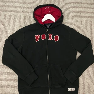 Svart hoodie från Polo Ralph Lauren med röd huva - Svart hoodie från Polo Ralph Lauren med röd insida på huvan och broderad text 'POLO' i rött framtill. Dragkedja hela vägen och fickor framtill. Märke och siffra 67 nertill. Tillverkad i bomull och polyester.