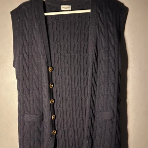 Mörkblå stickad väst från John Henric - Säljer en mörkblå stickad väst från John Henric med kabelstickat mönster. Västen har knappar framtill och två fickor. Perfekt att bära över en skjorta för en klassisk look. Använd en gång sedan blev den för liten. Inköpt för 650 kr på John Henric i Malmö. 