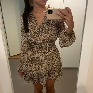 Leopardmönstrad kortklänning från Zara - Säljer en klänning från Zara med leopardmönster i beige och bruna toner. Klänningen har v-ringning, långa ärmar med resår och smock i midjan. Skriv för frågor!