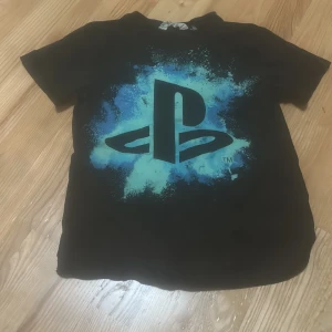 Svart PlayStation t-shirt från H&M - Cool svart t-shirt från H&M med stort PlayStation-tryck i blått och turkost på framsidan. Perfekt för dig som älskar gaming och vill ha en avslappnad stil. Klassisk passform och korta ärmar. Använd 1-4 gånger.