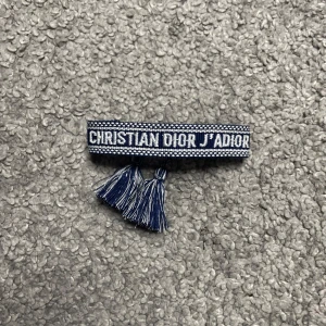 Dior armband med “J’ADIOR”-broderi (J’ADIOR Bracelet) – Blått - Dior armband med “J’ADIOR”-broderi (J’ADIOR Bracelet) – Blått  Säljer ett stilrent och lyxigt armband från Dior med den ikoniska “J’ADIOR”-texten broderad framtill! Köpt second hand men aldrig använt av mig – i princip i nyskick. Perfekt accessoar för dig som gillar designerplagg med en touch av bohemisk elegans.  ✅ Justerbar storlek med tofsar ✅ Vävd kvalitet och tydliga detaljer – talar för äkthet  • Färg: Marinblå med vit brodyr • Skick: Nyskick • Design: “J’ADIOR”-broderi med tofsar 127