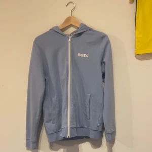 Ljusblå hoodie med dragkedja från BOSS - Säljer en ljusblå hoodie från BOSS med vit dragkedja och logga på bröstet. Tröjan har huva, två fickor framtill och ribbade muddar. Perfekt för en avslappnad stil.