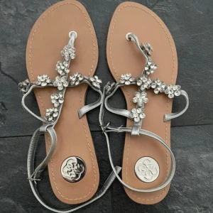Silvriga sandaler med blommiga strassdetaljer från Guess - Säljer ett par snygga sandaler från Guess i storlek 40. De har silvriga remmar och är dekorerade med blommor i strass. Perfekta för sommaren och ger en glittrig touch till din outfit. Platt sula och öppen tå. Helt nya, aldrig använda.