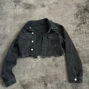Svart croppad jeansjacka - Säljer en svart croppad jeansjacka med råa kanter och klassisk krage. Jackan har långa ärmar, bröstfickor med lock och silverfärgade knappar framtill. Perfekt för dig som vill ha en edgy och trendig look.