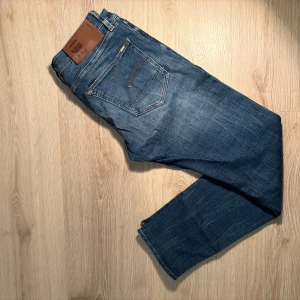 Blå jeansbyxor från G-Star - Säljer ett par klassiska blå jeans från G-Star med snygga slitningar och en bakficka med gul söm. Byxorna har normal passform och är tillverkade i jeansmaterial. Perfekta för en avslappnad stil.