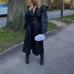 Svart skinn trenchcoat med bälte - Säljer en svart lång trenchcoat från NAKD i skinnimitation. Jackan har bred krage, långa ärmar och en cool oversized passform. Utan bälte denna på bilden tillhör en annan jacka.