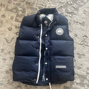 Mörkblå Canada Goose väst - Säljer en mörkblå dunväst från Canada Goose med vit logotyp på bröstet och tydlig märkning på fickan. 