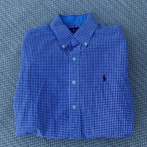 Blårutig skjorta från Ralph Lauren - Säljer en klassisk blårutig skjorta från Ralph Lauren som är helt ny endast prövad ett par gånger skjortan är i slim fit-modellen. Skjortan har långa ärmar, knappar framtill och är tillverkad i stretchig bomull. Perfekt för dig som gillar stilrena och tidlösa plagg. Perfekt skjorta inför sommaren. Tveka inte på att höra av dig vid frågor. Priset är ej spikat