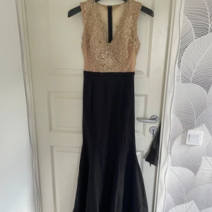 Svart och champagnefärgad långklänning med spetsdetaljer - Elegant långklänning med svart kjol och champagnefärgad överdel. Överdelen har v-ringning och är dekorerad med broderad spets och pärlor. Klänningen är ärmlös och har dragkedja i ryggen. Perfekt för bal eller fest.