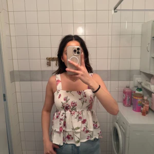 Blommig linnetopp från Story On - Supersöt blommig linnetopp från Story On i vitt med rosa och gröna blommor. Toppen har breda axelband, smockad överdel och volang nertill.🌸