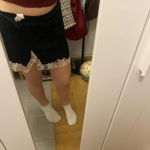 Svart kjol med spetskant - Säljer en svart kortkjol med dekorativ vit spetskant nertill. Kjolen har en omlott-look och är perfekt för dig som gillar en söt och trendig stil. Passar till många olika outfits och är enkel att matcha.