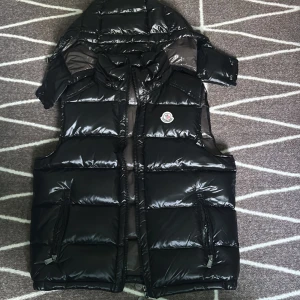 Svart dunväst från Moncler med huva - Säljer en svart glansig dunväst från Moncler med avtagbar huva och dragkedja framtill. Västen har två fickor med dragkedja och klassisk Moncler-logga på bröstet. Perfekt för kyliga dagar när du vill hålla stilen.