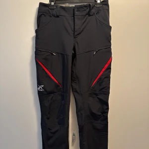 Svarta HiBall Pants från RVRC med röda detaljer - Säljer ett par svarta HiBall Pants från RVRC i storlek M. Byxorna har röda detaljer, dragkedjor vid fickorna och fleecefodrad insida för extra värme. Perfekta för utomhusaktiviteter och har en sportig look.