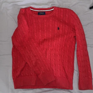 Röd stickad tröja från Polo Ralph Lauren - Säljer en röd stickad tröja från Polo Ralph Lauren med klassiskt kabelstickat mönster och broderad logga på bröstet. Tröjan har rund halsringning och långa ärmar. Perfekt för en stilren och tidlös look.