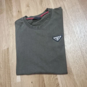 Prada T-shirt - Pris kan diskuteras                                          Säljer en olivgrön t-shirt från Prada med rund halsringning och korta ärmar. T-shirten har en liten trekantig logotyp på bröstet och en diskret röd detalj vid nederkanten. Perfekt för en stilren och avslappnad look.