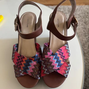 Klackskor strl 39 - Säljer ett par bruna sandaler med bred klack och öppen tå. Skorna har ett färgglatt, zickzack-mönstrat band i rosa, blått, rött och vitt över foten samt justerbart spänne vid ankeln. Perfekta för dig som vill sticka ut med färg och stil!