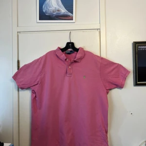 Rosa pikétröja från Polo Ralph Lauren - Säljer en klassisk rosa pikétröja från Polo Ralph Lauren i 100% bomull. Tröjan har korta ärmar, krage och knappar framtill samt den ikoniska lilla loggan broderad på bröstet. Perfekt för en avslappnad och stilren look.