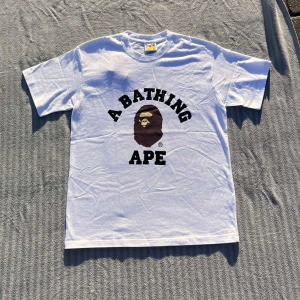 Vit t-shirt från BAPE med tryck - Säljer en vit t-shirt från BAPE med det klassiska 'A Bathing Ape'-trycket och apans ansikte på bröstet. T-shirten har rund hals och korta ärmar. Märkeslapp på ärmen och tillverkad i Japan. Perfekt för dig som gillar streetwear.