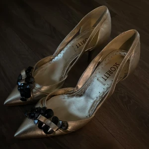 Guldiga pumps med svarta pärlor från Scorett - Säljer ett par eleganta guldiga pumps från Scorett med spetsig tå och dekorativa svarta pärlor och rosett framtill. Skorna har klack och är perfekta för dig som vill sticka ut med en glammig look. De är i storlek 37 men passar 37,5 med, klackar är burna en gång😊🌸