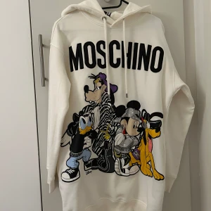 NY Vit hoodie med Disney-tryck från H&M x Moschino - Oversized fit. Säljer en vit hoodie från H&M x Moschino med stort tryck av Musse Pigg, Pluto, Långben och Kalle Anka på framsidan. Tröjan har huva med dragsko och ribbade muddar. Perfekt statement-plagg för Disneyfans!