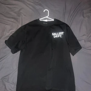 Svart t-shirt från Gallery Dept - Säljer en svart t-shirt från Gallery Dept. med vit text på bröstet och stor logga på ryggen. Klassisk passform och rund halsringning. Perfekt för dig som gillar streetwear och enkel stil.