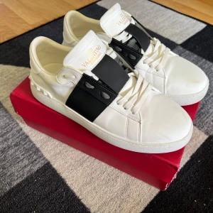 Valentino Open sneakers i vitt och svart - Säljer ett par snygga Valentino Open sneakers i vitt skinn med svart band över vristen. Klassisk rund tå och platt sula. Perfekta för dig som gillar stilrena och exklusiva sneakers med en modern touch. Bara lite smuts hittils på dem. Använt 5 ggr hittils och säljer därför dem e lite för stora. Inget slitage! Bra kvalitet, hör av er vid frågor och funderingar🔥🤝