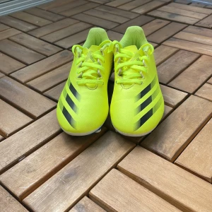 Neongula Adidas fotbollsskor - Säljer ett par neongula Adidas fotbollsskor med svarta ränder på sidorna. Skorna har snörning och är tillverkade i syntetmaterial. Perfekta för dig som vill sticka ut på planen!