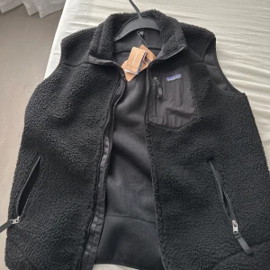 Svart fleeceväst från Patagonia - Svart fleeceväst från Patagonia med dragkedja framtill och två sidofickor med dragkedja. Västen har en bröstficka med dragkedja och mjuk, fluffig yta. Perfekt för lager-på-lager och svalare dagar. Tags kvar. 