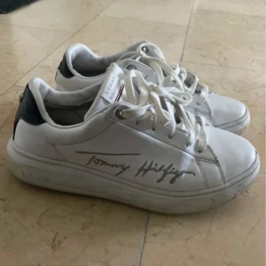 Vita sneakers från Tommy Hilfiger - Säljer ett par vita sneakers från Tommy Hilfiger med signaturdetalj på sidan och klassiska Hilfiger-färger på hälen. Skorna har snörning och rund tå, samt detaljer i mörkblått och rött baktill. Perfekta för en stilren och sportig look.Tommys Himfinger sneakers som blivit använda ett fåtal gånger och är därför i väldigt bra skick, dem säljes då dem är för små❤️