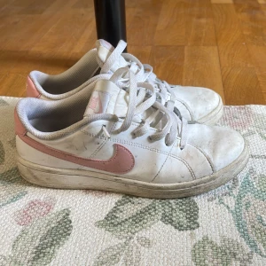 Vita och rosa Nike sneakers - Säljer ett par klassiska vita sneakers från Nike med rosa swoosh och häl. Skorna har snörning och rund tå, samt en platt sula. Perfekta för en avslappnad och sportig stil. lite smutsiga men annars väldigt fina.